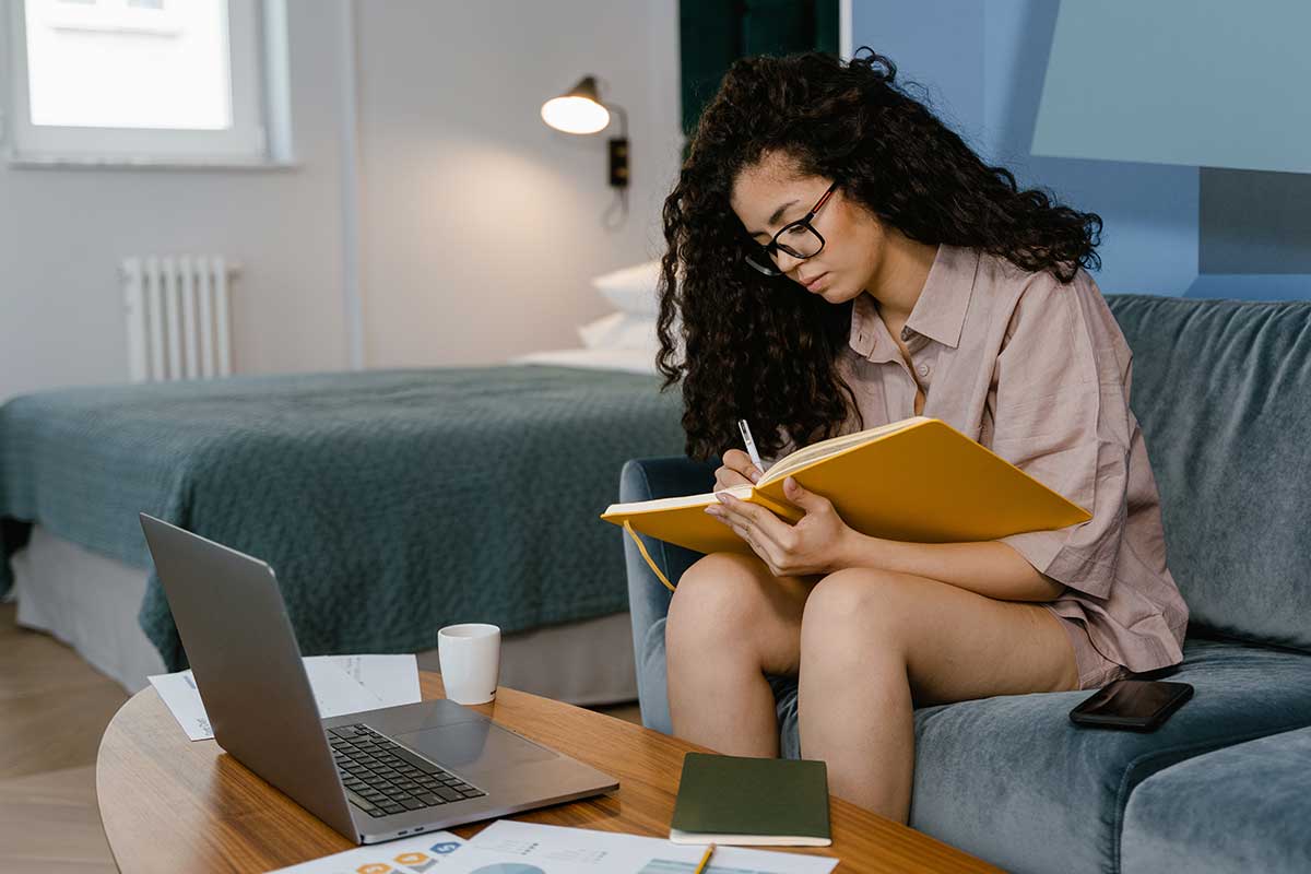 Kvinne skriver i bok sittende på sofa med laptop på bord foran