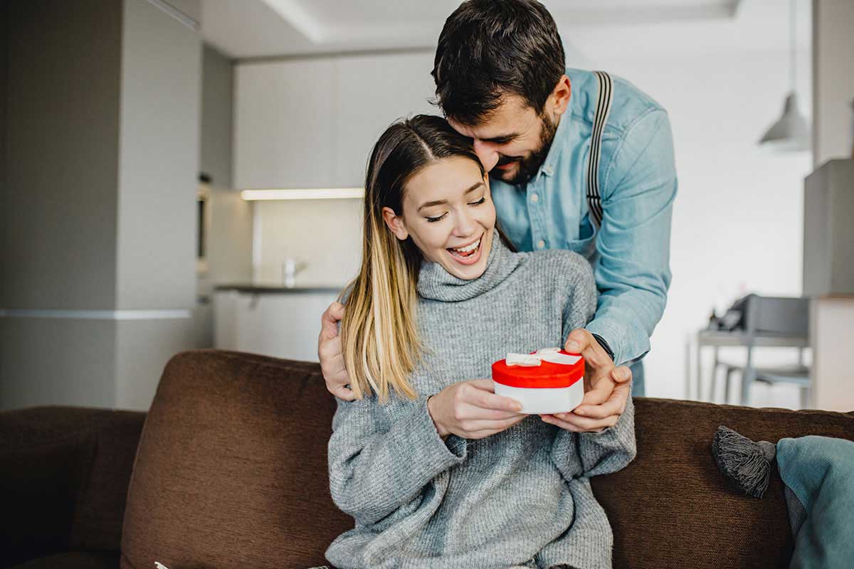 En mann lener seg over en kvinne som sitter i sofaen og gir henne en valentines gave som kommer i en boks med rødt lokk og hvit bunn.