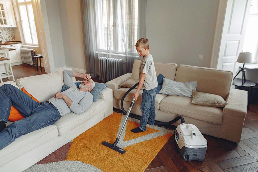 En mann som ligger utslitt på sofaen, mens et barn rydder og støvsuger hjemmet.